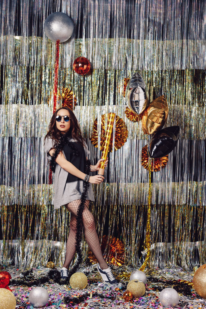 Happy young woman in fashionable clothes celebrating on a shimmer, colorful, party background. Party decorations gold and silver balls, balloons enjoying confetti, getting ready to open confetti.
