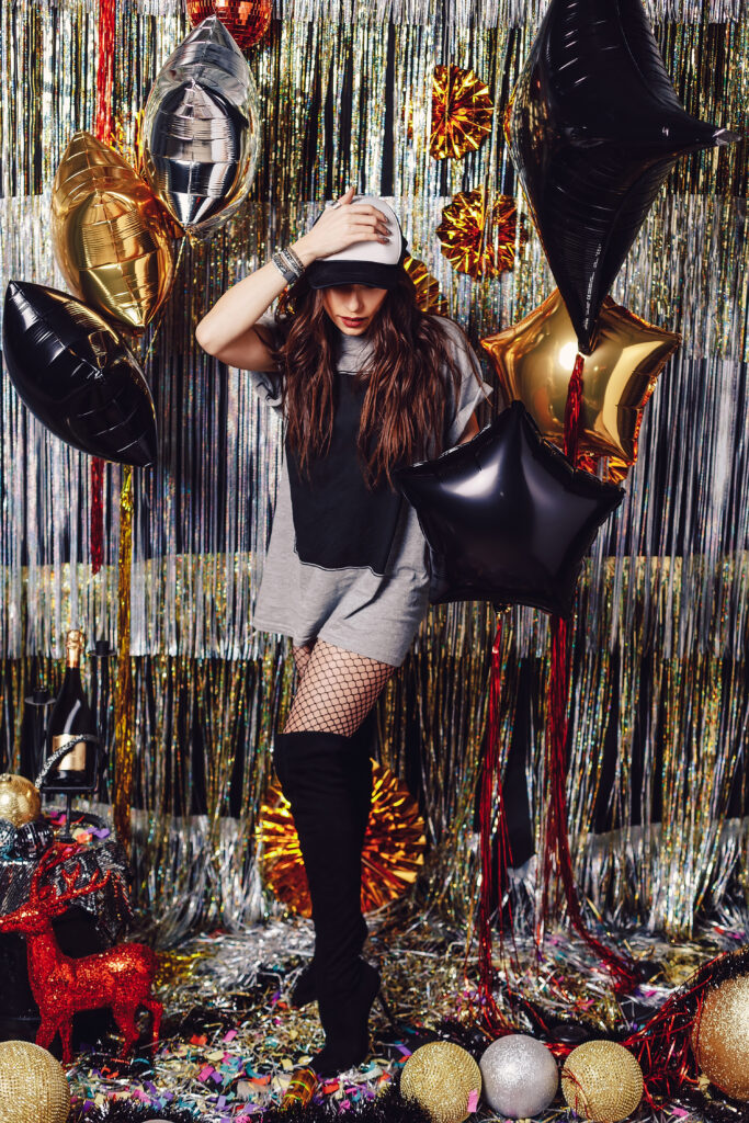 Young active dancing woman on a shimmer background. Party hard. Girl holding balloons, ready to celebrate in a space with golden and silver decorations. Sexy female with long high heels boots and cap.
